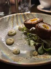 A close-up, eye-level shot of a Scotch egg starter served at a gastro pub. The dish is presented with a small side salad and dots of mustard mayonnaise on a textured, speckled plate.
