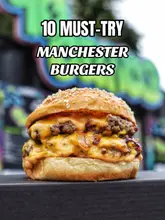 A promotional graphic featuring a close-up shot of a double cheeseburger with caramelized onions, overlaid with the text '10 MUST-TRY MANCHESTER BURGERS'. The image appears to be a digital creation or heavily processed, lacking the natural texture of a real food photograph.