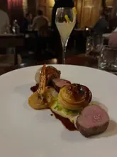 A well-composed, atmospheric shot of a fine dining main course, featuring pork belly and a pastry element, captured in a dimly lit restaurant setting. The shallow depth of field effectively isolates the food from the background.