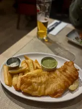 A standard, casual shot of a fish and chips meal at a gastropub. The image captures the meal and a drink in a typical pub setting, but lacks professional styling or careful framing.