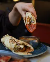 A close-up, high-contrast shot of a chicken pakora naan wrap being dipped into a vibrant red sauce, with a second wrap visible in the foreground. The lighting is dramatic, focusing attention on the texture and sauce of the wrap.