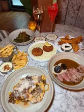 An overhead flat-lay shot of a table filled with a variety of small plates and two cocktails at a restaurant, showcasing a spread of food including steak, pasta, and fried chicken.