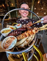 A person holds a large, tiered metal platter featuring a variety of Greek food, including skewers, sausages, pita bread, and dips, in a restaurant setting. The image is captured with a direct, harsh flash that creates significant overexposure on the food and the subject's face.