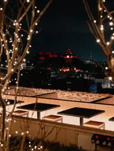 A high-quality, atmospheric night shot of a rooftop terrace in Edinburgh, featuring a clear view of the illuminated castle in the background. The composition uses foreground fairy lights to frame the scene, creating an inviting and premium feel for the venue.