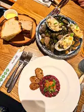 A high-quality overhead shot of a meal featuring beef tartare and tempura mussels, served with bread and butter. The presentation is clean and professional, highlighting the quality of the ingredients and the plating style of the venue.