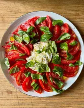 A vibrant, overhead food photography shot of a fresh tomato and mozzarella salad, featuring sliced heirloom-style tomatoes arranged in a circle with a central mound of creamy cheese, garnished with fresh basil, olive oil, and cracked black pepper on a rustic wooden surface.