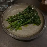 A top-down, moody shot of a plate of charred tenderstem broccoli, likely served as a side dish in a restaurant setting. The lighting is atmospheric and low-key, typical of a dinner environment, with the subject clearly defined against a dark table surface.