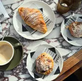 A high-quality, well-lit flat lay photograph of a cafe table featuring a croissant, an almond pastry, and a cup of coffee. The setting is clean and aesthetically pleasing, utilizing natural light to highlight the textures of the food and the marble tabletop.