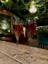 A close-up, eye-level shot of two glasses of rosé champagne placed on a wooden table, set against a dimly lit, atmospheric bar interior decorated with greenery and a chandelier.