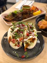 A close-up, high-angle shot of Indian-fusion bao buns topped with pomegranate seeds, red onion, cilantro, and sauces, served on a dark plate. In the background, other dishes like grilled meat are visible, suggesting a shared dining experience.