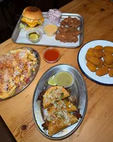 A top-down shot of a table spread featuring a burger, fried chicken, dirty fries, quesabirria tacos, and fried pickles at a casual restaurant.