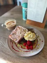 A close-up, well-lit photograph of a plate of pistachio French toast accompanied by a flat white, set on a wooden table.