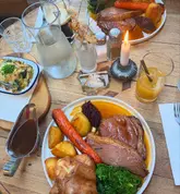 A high-quality, top-down shot of a Sunday roast spread at a pub, featuring a main plate with beef, roast potatoes, and vegetables, alongside a side of gratin and drinks.