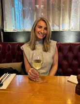 A portrait of a woman sitting at a restaurant table holding a glass of white wine, with a leather booth and window in the background.