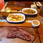 A top-down shot of a steak dinner at Flat Iron, featuring a flat iron steak, mac and cheese, and mash, with a cocktail in the foreground.