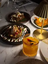 A high-quality, atmospheric shot of an Indian dining experience featuring small plates and a cocktail, illuminated by a warm table lamp.