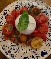 A top-down, close-up shot of a Burrata Caprese salad served on a blue-spotted ceramic plate. The image focuses on the central burrata ball surrounded by heritage tomatoes, capturing the freshness of the ingredients in a standard restaurant setting.