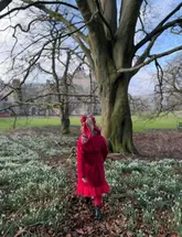 A photograph of a child in a red coat walking through a woodland area filled with snowdrops, with a large tree and a historic building in the background.