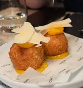 A close-up, eye-level shot of two Cacio e Pepe arancini balls, each topped with a dollop of egg yolk jam and shaved parmesan, served on branded paper.
