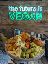 A close-up, user-generated style shot of a vegan Sunday roast carvery being held in front of a neon sign. The image captures the meal in a casual, authentic way, focusing on the food and the venue's branding.