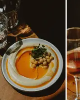 A high-quality, moody, and professional food photograph featuring a plate of hummus with chickpeas and parsley, accompanied by a glass of wine. The lighting is dramatic and intentional, highlighting the texture of the food.