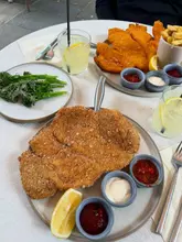 A top-down, well-lit shot of a table spread featuring crispy schnitzels, sides of broccoli and chips, and homemade lemonades.