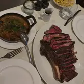 A top-down shot of a large, sliced ribeye steak served on a white platter, accompanied by a cast-iron skillet of mushrooms and artichokes, and a side of fries. The setting is a dimly lit, upscale restaurant environment.