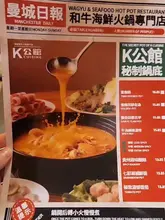 A close-up, slightly angled photograph of a printed restaurant menu for a hotpot venue, held by a hand. The menu displays various soup base options and pricing, with small, low-resolution images of the food.