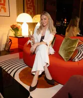 A portrait of a woman sitting on a bright orange sofa in a hotel suite, holding a glass of champagne. The setting features 1970s-inspired decor, including a saxophone, a large lamp, and bold artwork.