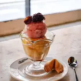 A close-up, eye-level shot of a lychee and mango sorbet dessert served in a glass, garnished with a cherry and accompanied by a fortune cookie on a branded plate.
