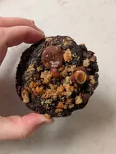 A close-up, high-angle shot of a homemade chocolate hazelnut cookie being held by a hand against a neutral, light-colored surface. The cookie features a dark, textured surface with visible chocolate chunks, hazelnut pieces, and a crumbly topping.