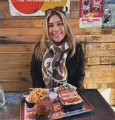 A casual, candid-style photo of a woman smiling at the camera, seated at a table with a metal tray containing a smash burger, fries, and patty melts. The setting appears to be a casual, rustic-style venue with wooden walls and posters.