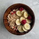 A simple, clean, overhead shot of a breakfast bowl featuring granola, sliced bananas, and raspberries in a wooden bowl.