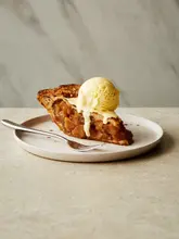 A professional, high-quality food photograph featuring a slice of apple pie topped with a scoop of melting vanilla ice cream, presented on a neutral, textured surface. The image is used to promote a baking cookbook.