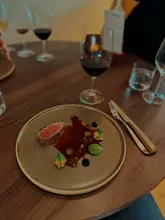 A high-quality, atmospheric shot of a fine-dining lamb dish, likely part of a tasting menu, set on a wooden table with wine glasses in the background. The lighting is warm and moody, characteristic of an upscale restaurant environment, highlighting the precision of the plating.