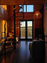 A beautifully composed, atmospheric shot of a wooden treehouse interior during golden hour. The image captures the warm, rustic aesthetic of the space, featuring a spiral staircase and a view through the glass doors to the outdoors, effectively conveying a sense of calm and seclusion.