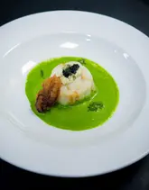 A close-up, high-angle shot of a fine dining fish dish featuring butter-poached Skrei cod, a tempura oyster, wild garlic foam, and caviar tartare, presented on a white plate against a dark, plain background.