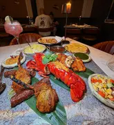 A high-angle shot of a lavish Indian feast, featuring a large platter with lobster, lamb chops, and chicken, accompanied by various side dishes and a cocktail, set on a marble table in a restaurant setting.