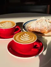 A bright, high-contrast photograph featuring two cups of latte with latte art and an almond croissant on a white table, capturing a sunny morning coffee break.