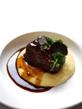 A high-quality, professional-looking food photograph of a braised short rib dish served on a bed of polenta, garnished with crispy kale and fresh truffle shavings. The image uses a clean, high-key aesthetic with a white background, emphasizing the rich, glossy texture of the meat and the elegant plating.