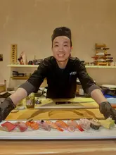 A medium shot of a sushi chef presenting a long, rectangular platter of various sushi pieces directly to the camera. The chef is smiling, wearing a black uniform, and standing behind a wooden sushi counter. The background shows a kitchen shelf with various bottles and kitchen tools.
