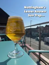 A bright, inviting shot of a glass of Menabrea lager placed on a teal table, with a blurred outdoor balcony background, effectively capturing the 'sun trap' atmosphere of the venue.