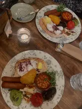 A top-down, overhead shot of two vegetarian breakfast plates served on floral-patterned plates, featuring poached eggs, avocado, hash browns, beans, grilled tomatoes, mushrooms, and halloumi, arranged on a wooden table with a lit candle.