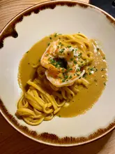 A top-down, overhead shot of a prawn pasta dish, featuring a single large butterflied prawn atop a bed of tagliatelle-style pasta coated in a bisque-like sauce, garnished with chives.
