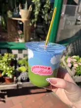 A close-up, eye-level shot of an iced matcha latte with cloud-shaped cold foam, held in a hand in front of a cafe storefront.