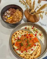 A bright, overhead food photography shot featuring a roasted carrot and dukkah flatbread alongside a chocolate-topped nice cream bowl, styled with a decorative vase on a clean white surface.