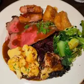 A top-down, flash-lit photograph of a Sunday roast dinner served at a restaurant. The plate features slices of beef, pigs in blankets, roast potatoes, cauliflower cheese, braised red cabbage, and greens, all covered in gravy.