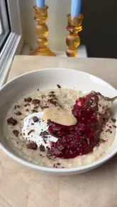 A short, top-down recipe video demonstrating the assembly of high-protein oatmeal topped with chia jam, peanut butter sauce, and chocolate shavings.