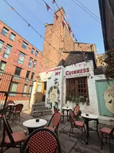 A bright, clear, and inviting wide-angle shot of a pub courtyard, featuring a distinctive 'Goodness My Guinness' mural on the brick wall and outdoor seating arrangements.
