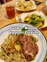 A top-down, overhead shot of a table spread featuring Uyghur cuisine, including a bowl of beef noodles, a plate of dumplings, a side of smashed cucumber, and a glass of tea. The image is taken from an overhead perspective, focusing on the main noodle dish.